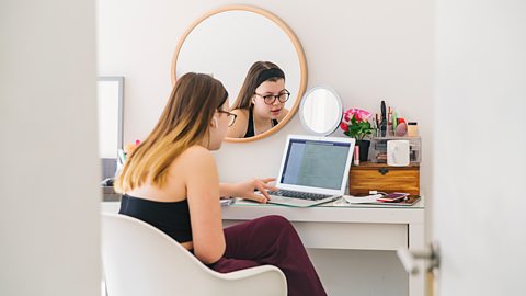 A girl sat doing work at her desk with a mirror hung above it 