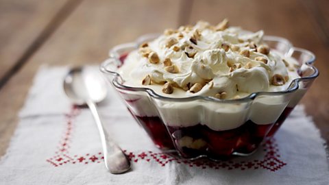 A glass serving bowl of trifle with silver spoon next to it. 