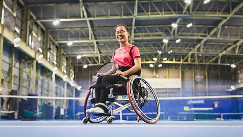 Tennis player in a wheelchair looking at the camera smiling. They are on a tennis court.