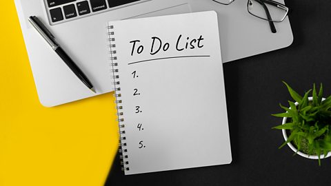 A 'to-do list' on a yellow and black desk. There are numbers 1 to 5 with no tasks listed next to them. The 'to-do list' is next to a laptop computer and pot plant.