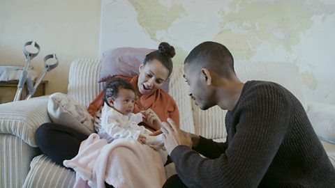 Mum, dad and their new baby sit on the sofa and have a play together. Mum is helping baby sit up.