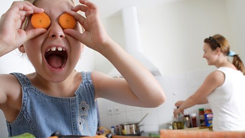 Girl plays with carrots while she cooks with her mum.