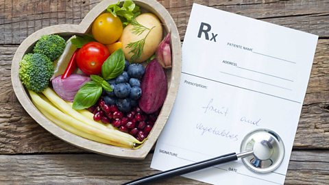 A wooden heart shaped bowl filled with fruit and veg with a doctor's prescription next to it prescribing fruit and vegetables