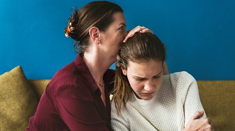 Mother comforts her daughter as they sit together on a sofa