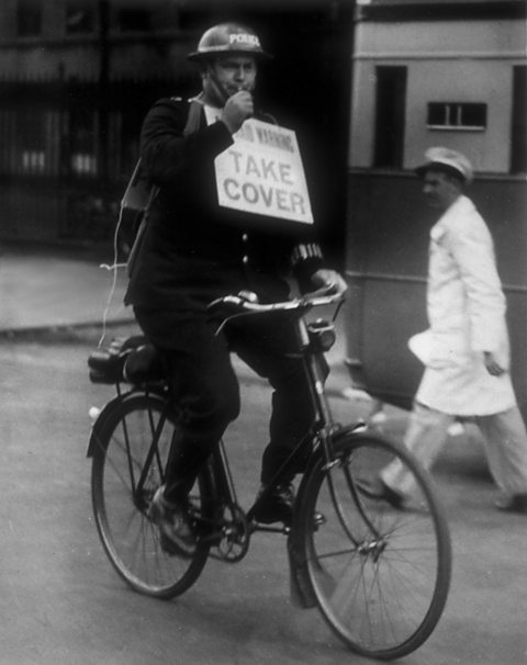 A policeman on a bicycle blows his whistle to warn the public of an imminent air raid.