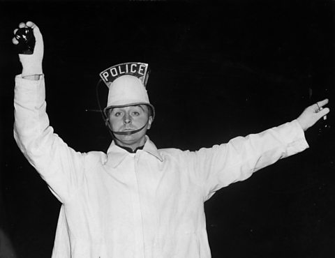 A policeman on point duty in the blackout wearing a long white coat and helmet with an illuminated sign saying 'Police'.