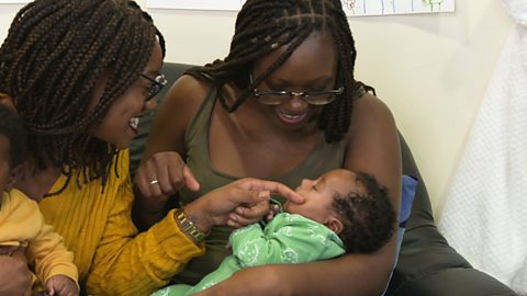 Two mums with their newborns sit on a sofa together, having a cuddle.