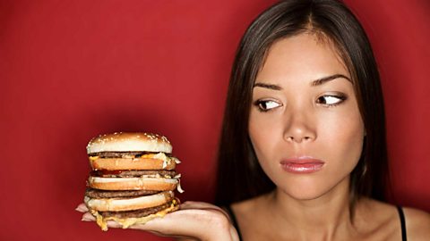 A woman holds a burger which has four patties and bread layers. She eyes it suspiciousy