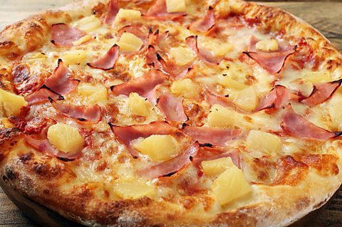 A thick-crust ham and pineapple pizza served on a table. The cheese on the crust of the pizza is golden brown and some of the edges of the ham are slightly blackened and charred.