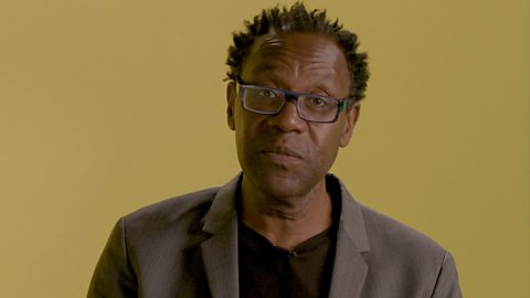 Sir Lenny Henry wearing a grey jacket standing in front of a mustard yellow background.