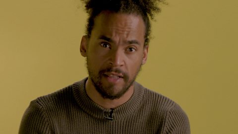 Dev Griffin wearing a grey jumper, standing in front of a mustard background.