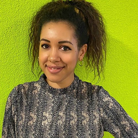 A photograph of Chloe Vethamony, teacher and Live Lessons presenter, wearing a grey top and smiling in front of a yellow wall.