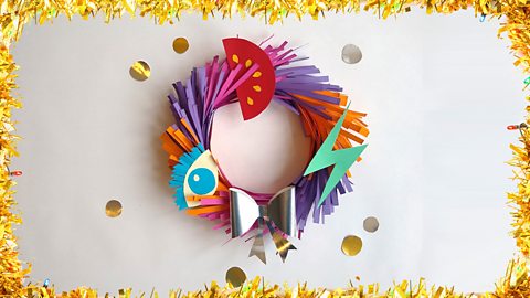 An alternative, colourful Christmas wreath with card cut-outs of a tomato, an eye, a lightning bolt and a silver bow decorating it.