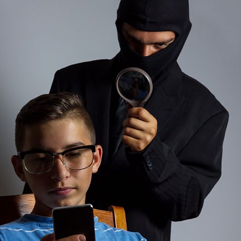 A person in a mask spying on a teen on his phone with a magnifying glass.