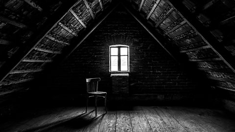 A black and white image of a sloped attic - an empty wooden chair sits in front of a small window.