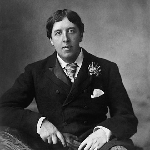 A photo of Oscar Wilde, sat in a suit with a carnation in his lapel.