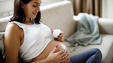 A pregnant woman sits on her sofa and massages lotion into her bump.