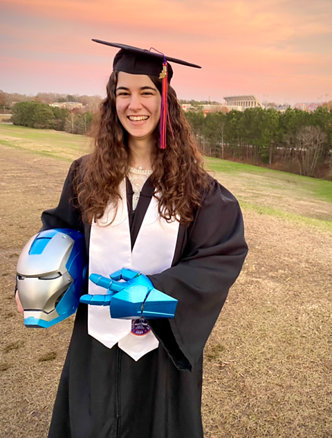 Emily Yarid wearing a graduation gown and holding a 3D printed blue and silver Iron Man helmet