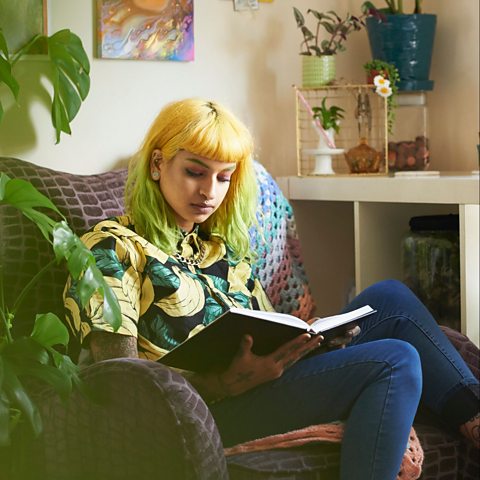 A girl with yellow and green hair sat on a comfy chair reading a book. There are plants on the cabinet behind her.