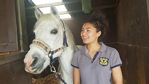 Mica Howard (right) standing next to a white horse (left) and looking off to the left of the frame with a beaming smile.