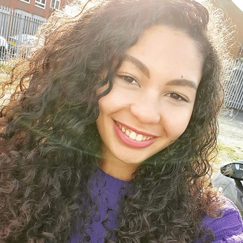 Mica Howard selfie smiling at the camera, she has light brown skin, brown eyes and lots of curly brown hair.