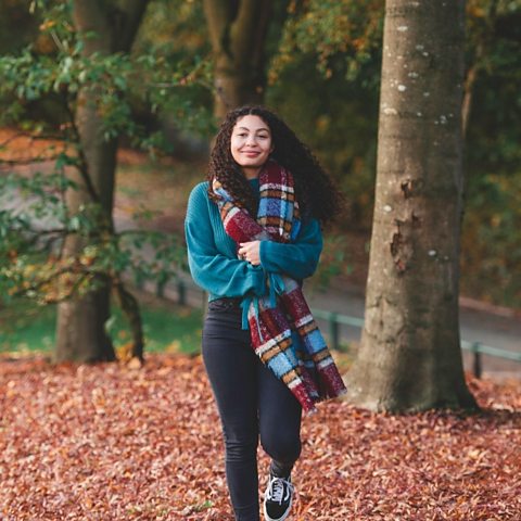 Mice is walking towards the camera smiling, she is wrapped in warm clothes and a scarf in an autumn woodland scene.