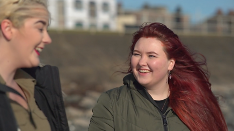 Amanda is walking on a beach, she is wearing a green coat and laughing with a friend.