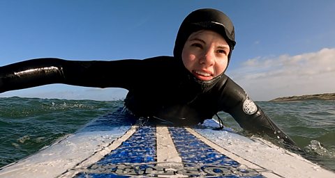 Hannah is wearing a black wet suite with hood up, she is on a blue and white surf board paddling on the water.