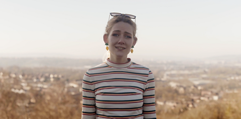 Chloe standing on a hill overlooking a town. She is wearing a white and orange striped sweater and sunglasses on her head.