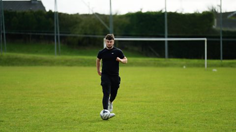 Adam is wearing a black teeshirt and jogging bottoms. He is playing football on a grass pitch, there is a goal behind him.