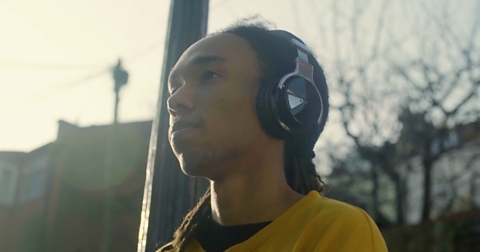 Kaden listening to music on headphones, he is walking down an urban street, wearing a yellow jumper.