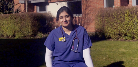 Samya sitting in a garden, she is wearing a blue medical outfit with a scethascope around her neck. 