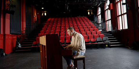 Caothan sitting on a piano. They are on a stage in front of red theatre seating.