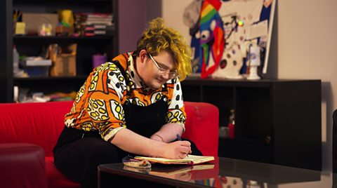 Kit is sitting on a red sofa drawing in a notepad, he is wearing black dungarees and an orange shirt with patterns on it.