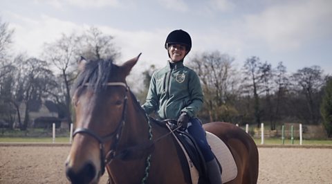 Tegan is in a menage, she is sitting on a brown horse and wearing a green jacket and black riding helmet.