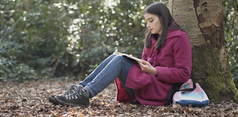 Amy sitting next to a tree reading a book. She is wearing a purple coat and jeans.