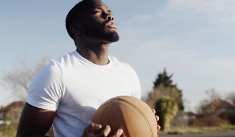 Donnell wearing a white teeshirt holding a basketball. He is outside near some trees.