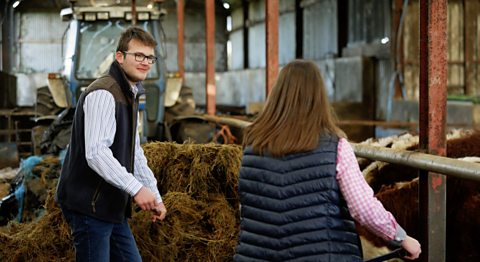 Nathan is wearing a green gilet and white shirt. He is in a barn near some hay bales and a tractor looking at someone.