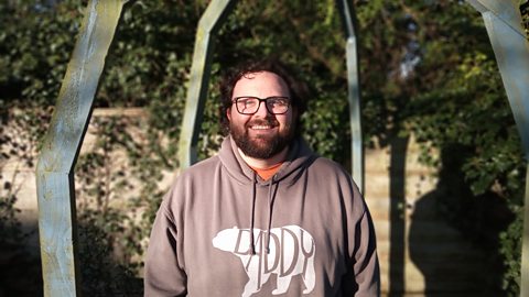 An adult male - Greg Baldwin - standing outside, smiling at the camera and wearing a brown hoodie. 