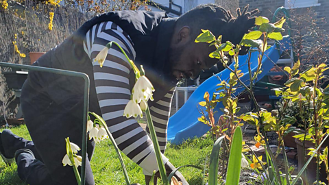 Image of Raoul Lindsay kneeling on the ground and gardening. He is wearing a striped jumper and is surrounded by green plants.