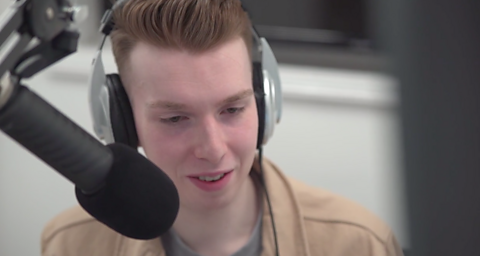 Alex is in a radio studio next to a microphone. He is wearing a brown jacket and silver headphones.