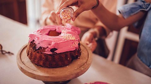Person pouring colourful sprinkles onto a donut shaped cake which is on top of a wooden cake stand on a work surface.
