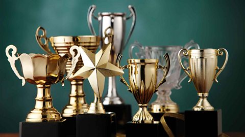 Seven trophies, five are gold, one silver and one glass. One trophy is shaped like a star, the rest are traditional trophy shaped.