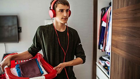 Student wearing a black t-shirt and red headphones holding a red laundry basket full of clothes.