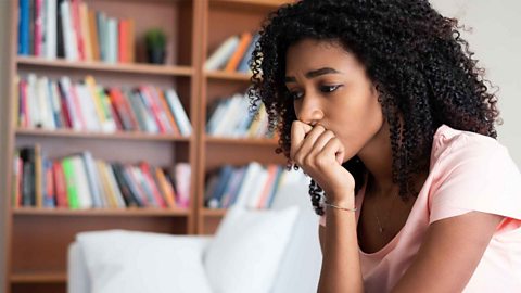 A young girl looking worried. She is wearing a pink t-shirt sitting on a white sofa in front of a book case full of books.