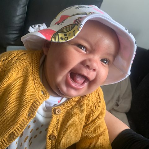 A very cute picture of baby Delilah, wearing a yellow cardigan and a hat, smiling.