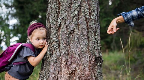 Photograph of a small blonde girl wearing a pink hairband and rucksack, with her ear to a tree. On the other side of the tree, a hand is about to knock on the tree trunk.