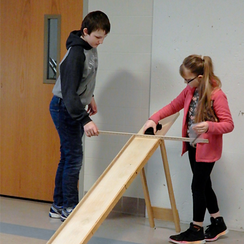 Two students watch as a car rolls down a ramp.