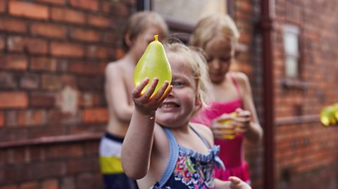 A young female child is standing outside in swimming costume and is holding a water balloon in the air, looking at it and smiling. There are children out of focus in the background playing with water balloons.