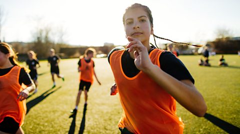 A young woman/teenager is mid run on a field. She is wearing a bright orange vest. In the background other women can be seen running on a field in vests.